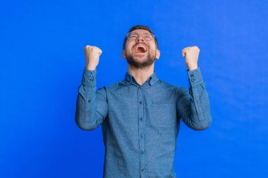 Mutlu beyaz adam başarıyı kutluyor zaferi, doğum gününü, piyango hedefini başarıyı iyi haber zaferini. Genç yakışıklı adam mavi stüdyo geçmişinde izole edilmiş.