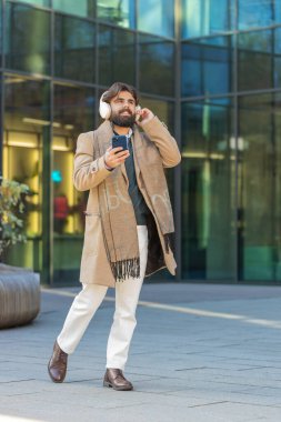 Mutlu Arap işadamı kablosuz kulaklık takıyor, açık havada akıllı telefondan en sevilen enerjik müziği dinliyor. Gülümseyen adam şehir sokağının yanından geçiyor. Yaşam Tarzları.