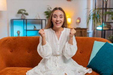 Mutlu genç bir kadın TV izlerken bağırıyor, başarıyı kutluyor, kazanıyor, gol başarısı iyi haber, piyango şansı, oturma odasında koltukta oturan zafer. Evde beyaz elbiseli bir kız var. Yaşam biçimi