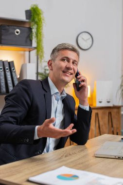 Genç işadamı bilgisayarını kullanıyor ve cep telefonuyla müşteriye cevap veriyor. Resmi takım elbiseli beyaz adam uzaktan kumanda işinde çalışıyor, ofis masasında konuşuyor..