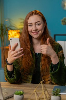 Çevrimiçi akıllı telefon uygulaması kullanarak gülümseyen mutlu kadın ev ofisinden çalışan uzaktan kumandayı işaret ediyor. Serbest çalışan kız iyi bir mobil program kullanmayı öneriyor. Başparmaklar yukarı.