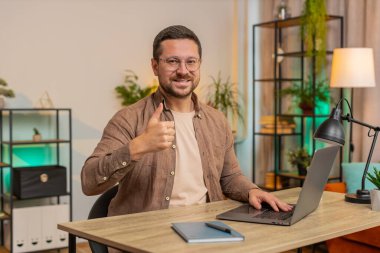 Gibi. Laptop kullanan mutlu, genç, beyaz bir adam kameraya bakıp olumlu işaret veriyor. İyi haber, olumlu geri bildirim. İş adamı ofis masasında oturuyor.