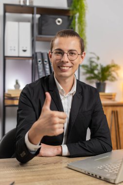 Mutlu, aşırı neşeli, heyecanlı, genç, Kafkasyalı adam. İnternetteki zafer konseptini kutlamak için dizüstü bilgisayarla heyecanlı. İç işlerindeki masada oturan çifte başparmakları gösteren kameraya bakıyor. Takım elbiseli adam.