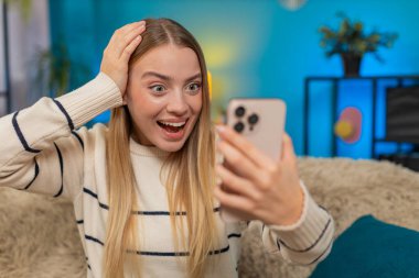 Beyaz bir kadın evdeki kanepede oturmuş online oyunu kutluyor ve zaferi kazanıyor. Heyecanlı kız akıllı telefonu tutuyor, sevinçle el kaldırıyor, gülümsüyor, mutluluğu, heyecanı ve başarıyı içeride hissediyor.