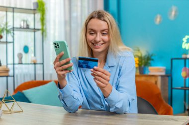 Kredi kartı ve akıllı telefon kullanan genç bir kadın para transferi yaparken internetten alışveriş yapıyor ev ofisinin masasında oturuyor. Çevrimiçi uzaktan kumanda işinde çalışan neşeli bir kız..