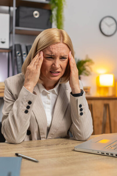 Усталая взрослая деловая женщина чувствует головную боль, работая за ноутбуком в домашнем офисе. Грустная перегруженная женщина-фрилансер в формальном костюме массирует виски и массирует больное место руками.