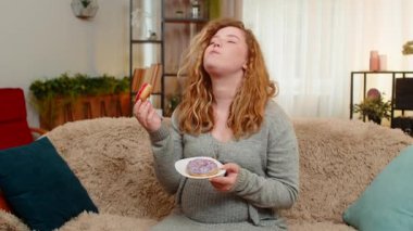 Hamile, genç, mutlu bir kadın elinde donutla kanepede oturuyor. Neşeli bir ifadeyle gülüyor, tatlı bir zevkin tadını çıkarıyor. Kız küçük zevkleri kucaklıyor, mutluluğu basit ve lezzetli anlarda buluyor..