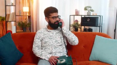 Hintli adam evinde koltukta oturup 90 'lardan kalma antika bir telefondan heyecanlı bir şekilde iyi haberler dinliyor. Arap adam, elinde bir cihaz tutarken sevinç ve ilgi gösteriyor ve içeride gülümsüyor..