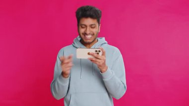 Bağımlı genç Hintli adam cep telefonuyla yarış oyunları oynuyor. Pembe stüdyo arka planında izole edilmiş zaferi kutlayan sürücü simülatörlü akıllı telefon uygulaması kullanan adam..