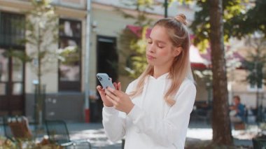 Mutlu gülümseyen beyaz kadın akıllı telefon kullanıyor internette mesajlar yazıyor, işi bitiriyor, dışarıdaki kameralara bakıyor. Şehir caddesinde duran ergen kız. Kasaba yaşam tarzları.
