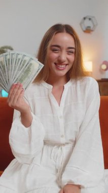 Başarılı beyaz kadın elinde dolar banknotları tutan bir hayranla sınıftaki kanepede oturuyor. Mutlu heyecanlı yetişkin kız çevrimiçi piyango oyunu kazanarak tatil planlama bütçesi hesaplıyor. Dikey.