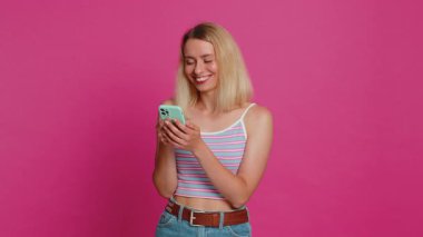 Beyaz bir kadının mesajlaşması akıllı telefon sosyal medya uygulamalarında içerik paylaşımı, rahatlama filmi izlemesi. Genç kız pembe arka planda izole edilmiş cep telefonu gülümsemesi kullanıyor.