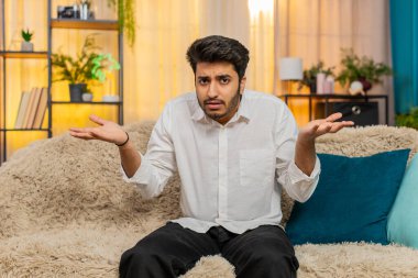 Hintli adam şaşırmış görünüyor, el kaldırıyor ve kafa karışıklığını dile getiriyor. Arap adam kanepede oturuyor, inanamıyor, şaşırıyor ve beklenmedik bir şeyi sorguluyor.