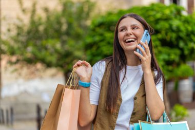 Alışveriş sonrası alışveriş torbaları açık havada satılan mutlu gülümseyen beyaz kadın. Kız turist şehir caddesinde durmuş akıllı telefondan uzaktan konuşma yapıyor..