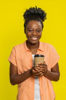 Afro-Amerikalı bir kadın elinde bir fincan plastik kahve tutuyor, aroması ve tadı ile mutlu ve tatmin olmuş bir bakışla. Sarı arka planda izole edilmiş siyah bir kız. Keyif verici. En sevilen sabah içme ritüelinden.