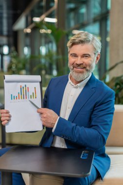 Olgun Kafkasyalı patron iş adamı çevrimiçi video sunumu yapıyor, grafik analizleri gösteriyor, planlama bütçesi, istatistik. Ofis lobisinde yakışıklı bir adam kameraya bakıyor, konuşuyor..