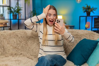 Beyaz bir kadın evdeki kanepede oturmuş online oyunu kutluyor ve zaferi kazanıyor. Heyecanlı kız akıllı telefonu tutuyor, sevinçle el kaldırıyor, gülümsüyor, mutluluğu, heyecanı ve başarıyı içeride hissediyor.