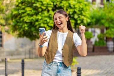 Kafkasyalı genç bir kadın cep telefonuyla kutlama yapıyor iyi haber kazanıyor, piyango büyük ikramiyesi kazanıyor, açık havada dağıtıyor. Şehir caddesinde duran mutlu yetişkin bayan turist. Kasaba yaşam tarzları.