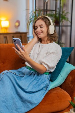 Kablosuz kulaklıklarla mutlu genç bir kadın evinde koltukta oturup en sevdiği enerjik disko dans müziğini dinliyor. İnsanlar hafta sonları boş zaman aktiviteleri yapar. Dikey.