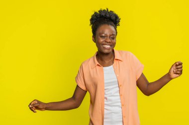 Afrika kökenli Amerikalı bir kadın komik el hareketleriyle dans ediyor, neşe ve neşe saçıyor. Sarı arka planda müzik dinleyen, neşeli ritim ve kaygısız tavır sergileyen siyah bir kız.