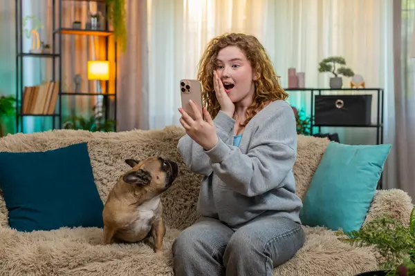 Beyaz saçlı genç bir kadın akıllı telefondan harika haberler okurken kanepesinde sevinç ve heyecanla gülümsüyor. Yanında pug dog olan bir kız var. İkisi de pozitif enerjiyle tepki veriyor..