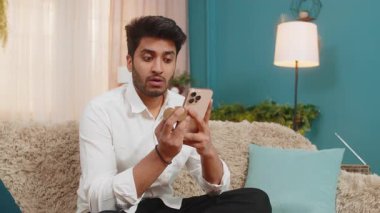 Kanepede oturan Hintli adam akıllı telefonda bitcoin cüzdan bakiyesini kontrol ediyor, biraz gergin hissediyor. Adam kripto döviz piyasasındaki dalgalanmalara odaklanmış. Gerçek zamanlı finansal değişimlere tepki veriyor.