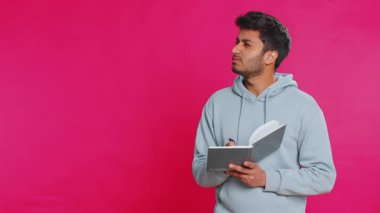Düşünceli gazeteci genç Hintli adam notlar alıyor, not defteri günlüğüne kalemle düşünceler yazıyor, liste yapmak için, iyi fikir. Arap Hindu adam pembe arka planda izole edilmiş.