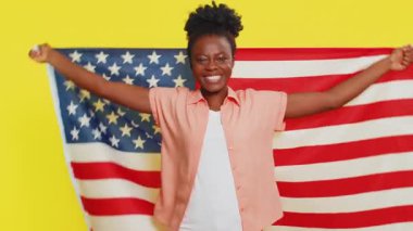 Amerikan bayrağını sallayıp saran mutlu Afrikalı Amerikalı kadın insan haklarını ve özgürlüklerini kutluyor. Bağımsızlık Günü. Başkanlık seçimi. Sarı arka planda izole edilmiş genç siyah kız.