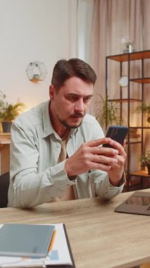 Mutlu genç adam akıllı telefon daktilo kullanarak evet diyor, başarıyı kutluyor, piyango hedefini kazanıyor olumlu haberler, zafer. Evdeki adam masada oturuyor. Dikey.