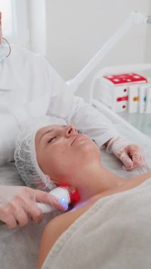 Kozmetik uzmanı, kadını yüz ve boyun bölgesinin kaldırma prosedürü, kolajen maskesi yapar. Güzellik uzmanı vakumlu yüz masajı terapisi için elektrikli ekipman kullanıyor. Cilt bakımı, güzellik tedavisi. Dikey çekim