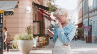 Kafası karışmış genç bir kadın akıllı telefon kullanıyor sosyal medya mesajları yazıyor açık hava gezici uygulamasında haritada bir yol arıyor. Şehir caddesinde duran gözlüklü dalgın gezgin..