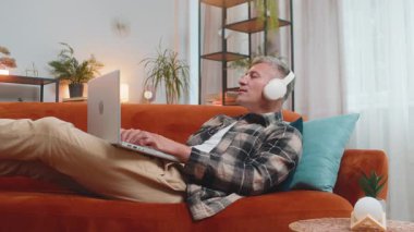 Kablosuz kulaklıklı, evinde kanepeye uzanmış, dizüstü bilgisayar ağı üzerinde çalışırken disko müziği dinlemeyi seçen mutlu, rahat, beyaz bir genç adam. Erkekler hafta sonları eğleniyor.