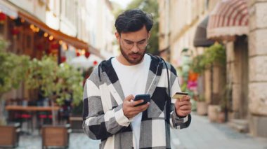 Hintli genç adam para transfer ederken kredi kartı akıllı telefonu kullanıyor, online alışveriş yapıyor, açık havada yemek dağıtım odası sipariş ediyor. Şehir caddesinde gülümseyen İspanyol adam.