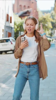 Kablosuz kulaklıklarla gülümseyen, dans eden, favori enerjik disko müziğini dinleyen mutlu genç kadın akıllı telefonları açık havada zaferi kutluyor. Şehir caddesinde duran kız. Kasaba yaşam tarzları.