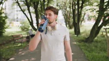 Sportif atletik susamış beyaz erkek koşucu parkta çalıştıktan sonra su içiyor, spor şişesi içiyor aktif genç yaşam tarzı ferahlatıcı su içiyor gülümseyerek uzaklaşıyor..