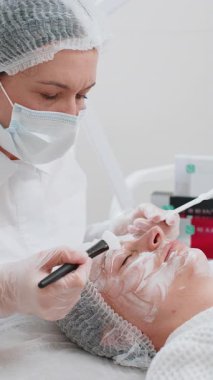 Güzellik uzmanı, genç bir kadının yüzüne fırçayla nemlendirici kimyasal krem maske takıyor. Kaplıcadaki kız cilt bakımı. Yüz kozmetolojisi tedavisi. Enzim terapisi. Dikey