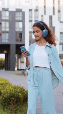 Kablosuz kulaklıklı, neşeli, çocukluk çağındaki mutlu çocuk dışarıda akıllı telefon dansında en sevilen disko rock n roll müziğini dinliyor. Şehir caddesinde yürüyen bir kız. Dikey