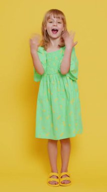 Genç sarışın kız kameraya gülümseyip el sallayarak merhaba ya da hoşça kal diyor, misafirperver bir ifadeyle karşılıyor. Sarı arka planda izole edilmiş genç kız çocuk. Dikey