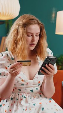 Genç kızıl saçlı kadın kredi kartı ve akıllı telefon kullanarak para transfer ediyor, internetten nakit para almıyor, evde yemek siparişi veriyor. Mutlu kız kanepede oturuyor. Dikey