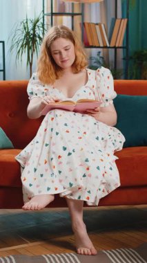 Mutlu kızıl saçlı kadın ilginç kitaplar okuyor, sayfaları çeviriyor edebiyatın tadını çıkarıyor, rahat bir koltukta dinleniyor. Evdeki kanepede oturan kızıl saçlı kız. Dikey