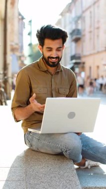 Şehir sokağında oturan Hintli serbest çalışan, laptop kamerasına bakan, arkadaşlarıyla, ailesiyle ya da müşterisiyle görüntülü konuşma yapan, uzak sohbetlerden hoşlanan. El sallayan adam. Dikey