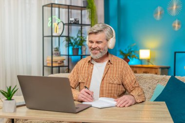 Beyaz, olgun bir adam evde oturmuş kulaklıklı bir dizüstü bilgisayardan podcast ya da online yayın dinliyor. Adam not defterine not tutuyor, içerikle ilgileniyor, eğitimi öğreniyor.