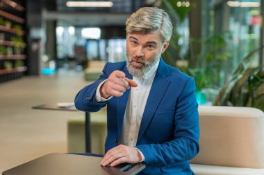 Seni seçiyorum. Kafkasyalı orta yaşlı işadamı kamerayı işaret ederek, mutlu bir ifadeyle bakarak, işe alma seçimini yapıyor. Resmi takım elbiseli mutlu adam modern ofis otel lobisinde oturuyor..