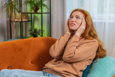 Hasta kızıl saçlı kadın baş ağrısı problemini tedavi etmek için tapınakları ovuyor, tansiyon, migren, stres, ekşitme, evde yüksek tansiyon. Genç yetişkin bir kız odada kanepede..