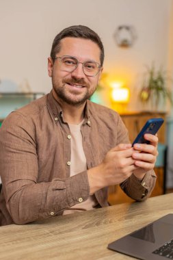 Masada oturan, cep telefonunu kullanan ve gülümseyen genç, beyaz bir adam. Guy Freelancer akıllı telefon sosyal medya uygulamaları üzerinden online film izlerken mesajlarını paylaşıyor.