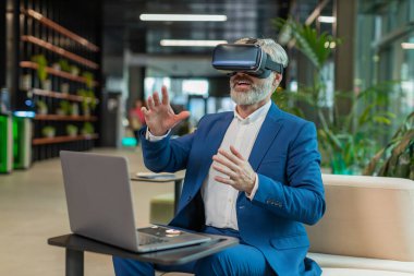 Olgun bir iş adamı yazılım geliştiricisi kulaklık uygulaması kullanarak sunum yapıyor. Sanal gerçeklik 3D 360 videosu izliyoruz. Ofis lobisinde çalışan, VR gözlüklü, beyaz saçlı, takım elbiseli adam..