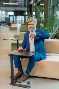 Hoşlanmamak. Takım elbiseli üzgün işadamı ofis lobisinde oturmuş işaret dilini gösteriyor hoşnutsuzluğunu, memnuniyetsizliğini ve kötü işini dile getiriyor. Otel odasında ciddi bir olgun adam var..