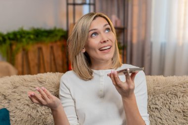 Orta yaşlı bir kadın evdeki koltukta akıllı telefon hoparlörü kullanarak bir şeyleri açıklamaya çalışıyor. Yetişkin kadın duygularını odaklanmış ve tutkulu bir şekilde ifade ediyor. Sözlü enerji el hareketiyle tepki veriyor..