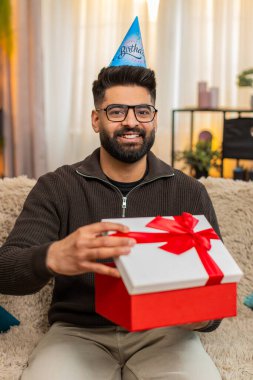 Evdeki Hintli adam mutlu bir şekilde doğum günü hediyesini açarken koltukta gülümseyerek ve heyecanla oturuyor. Arap adam şaşırmış ve neşeli görünüyor. Beklenmedik bir hediye ve özel bir kutlama anı karşısında heyecanlanıyor.