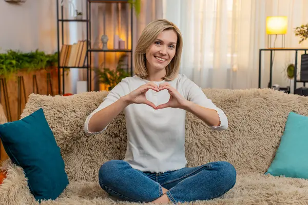 Orta yaşlı, neşeli bir kadın, elleri önde, kanepede oturmuş nazikçe gülümserken kalp şeklinde şekiller oluşturur. Yetişkin kadın sembolik ve duygusal jestlerle sevgi, sıcaklık ve şefkat gösteriyor.